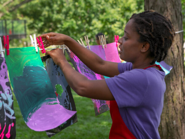 Girl in purple shirt and red apron hanging an anstract artwork on a string tied to a tree in the Art Garden