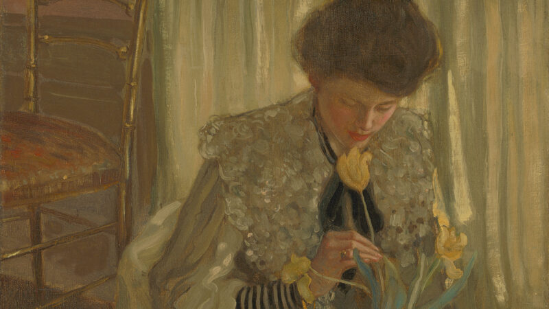 A young woman is seated on the floor leaning forward to admire one of three yellow tulips that extend from a blue and white Delftware bulb vase.