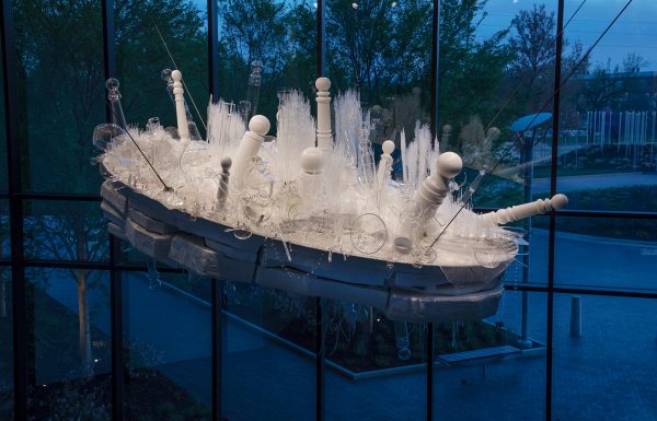 An overhead suspended tilted oval white and clear sculpture with top layer filled with assorted painted wood and glass barber poles, grass, chains and vessels, a middle white oval flint rock shapes and downward pointing painted wooden branches and poles underneath.