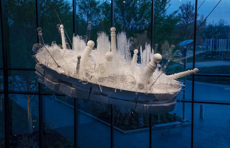 An overhead suspended tilted oval white and clear sculpture with top layer filled with assorted painted wood and glass barber poles, grass, chains and vessels, a middle white oval flint rock shapes and downward pointing painted wooden branches and poles underneath.