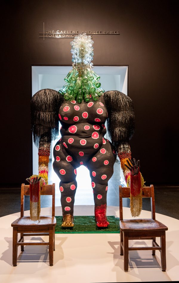Curvaceous black female figure with pink spots, glass coke bottled head, with outstretched beaded striped arms resting on two children's chairs.