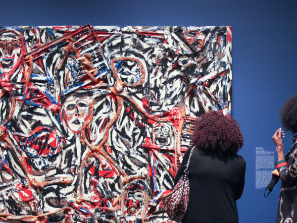 Two black women, one in a black dress carrying a leopard purse and the other searing a black dress with red lines and white, circular shapes, stand in front of a rectangular abstract artwork painted with the colors white, red, black, blue and dark orange, hung against a blue wall