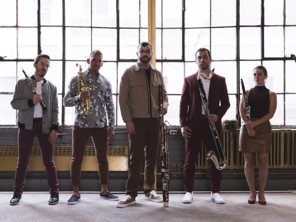 Group of people holding musical instruments standing in front of a bank of tall windows