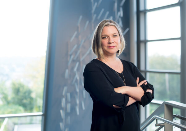 White woman standing with arms crossed. She has medium length blonde hair and is dressed in black with a gold necklace. She stands in front of a window and the wall behind her has a reflection of a geometic pattern.