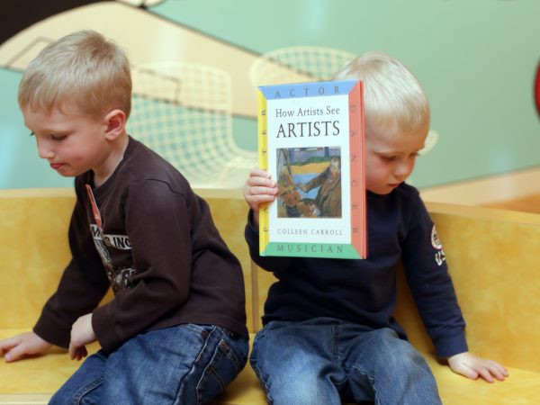 Two little boys who are white with blonde hair sit on a wooden bench facing away from each other while one of them holds up a book. They have on dark long-sleeve shirts and jeans.