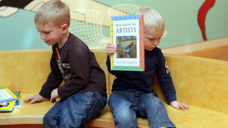 Two little boys who are white with blonde hair sit on a wooden bench facing away from each other while one of them holds up a book. They have on dark long-sleeve shirts and jeans.