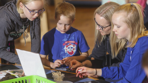 Three children and an adult, sitting at a talbe with art supplies in front of them. Children and adult are all blonde, wearing blue shirts. the girl holds a crayon.