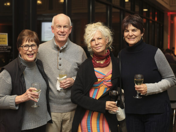Four people standing arm in arm and holding drinks. Woman on far left is dressed in a gray shirt and black vest and has short brown hair. Man is dressed in gray shirt and kakhi slacks and has a fringe of whtie hair. Woman in center right has tousled white hair and is dressed in a rainbow striped top with a black shrug sweater and bright red scarf around her neck. The woman on the left is dressed in black and has short dark hair.