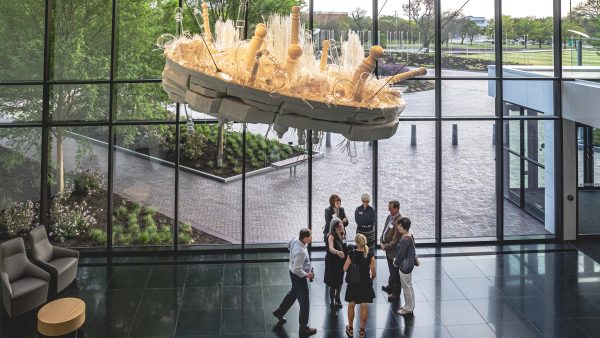 Group of 7 people standing in a semicircle below a hanging sculpture of clear glass objects and wooden poles on a white wooden base by artist Beth Lipman. The sculpture hangs in front of a wall of windows and overlooking a red brick driveway and walkway. The floor is polished black granite.
