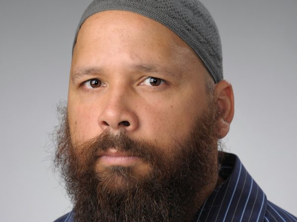 Photo of a Black man with a mustache and goatee wearing a grey head covering and a blue striped collared shirt