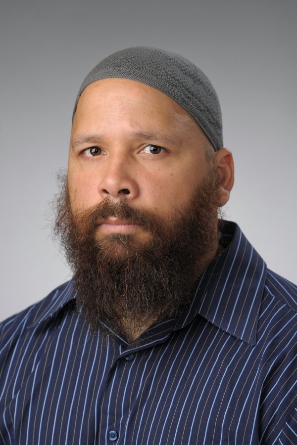 Photo of a Black man with a mustache and goatee wearing a grey head covering and a blue striped collared shirt
