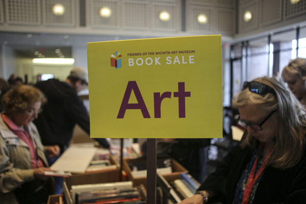 ellow sign in the foreground with text Friends on Wichita Art Museum Book Sale, ART on a stick above boxes of books on a table. Two women in either side of the table looking at the boxes of books.