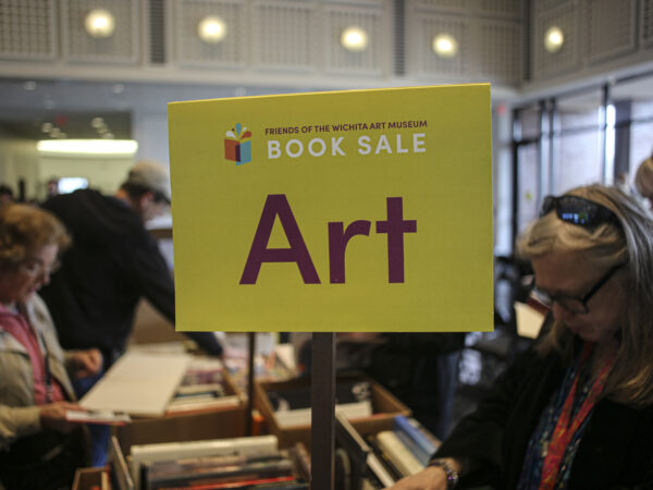 ellow sign in the foreground with text Friends on Wichita Art Museum Book Sale, ART on a stick above boxes of books on a table. Two women in either side of the table looking at the boxes of books.