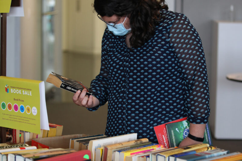 Woman in face mark holding a paperback book in her hand, standing at a table of books.
