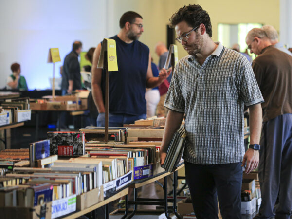Man with dark hair and weaing a light-colored plaid button-down shirt staning in fron of a table covered with boxes of books.