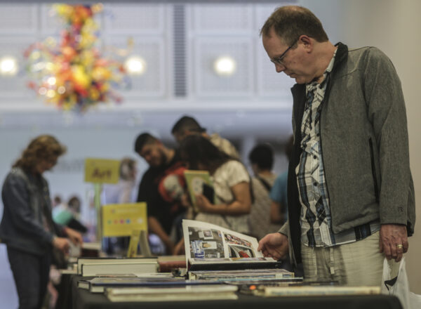Man dressed in a dark jacket and light slacks, standing at a table with books, turning a page in one of the books.