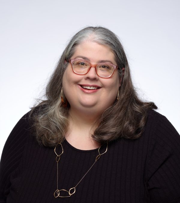A white woman with glasses and long hair wearing a black shirt and a necklace smiles for the camera