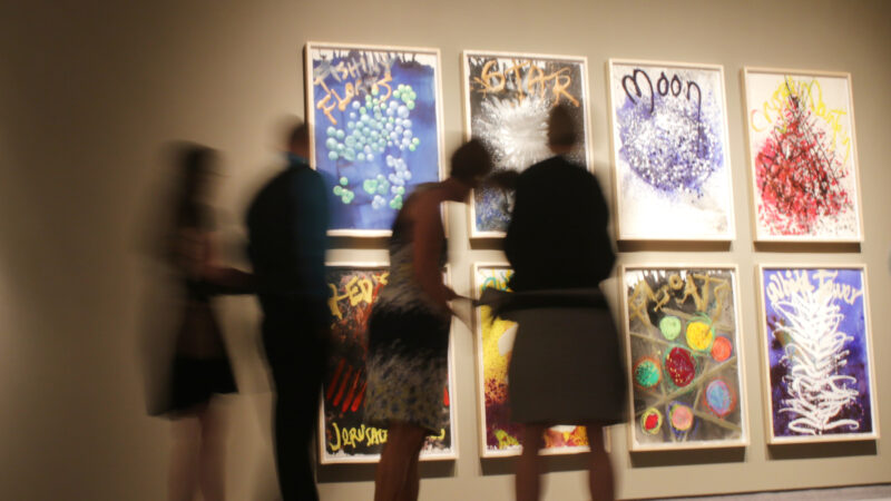 Photo of visitors blurry in the foreground and eight colorful prints on the wall in the background stacked in two rows and four columns