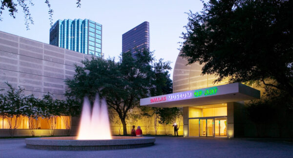 Photo of the exterior of a building with two people sitting on the edge of the fountain out front at dusk with a photographer taking their photo as the flash goes off