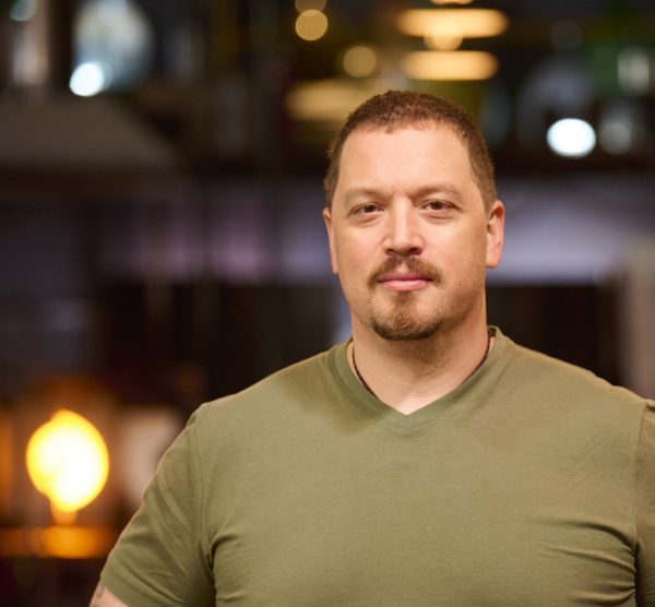 Photograph of a man with red hair and beard, dressed in an olive green shirt, with a dark background