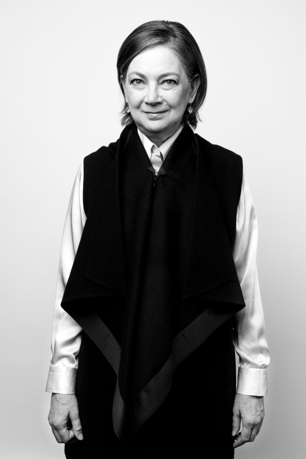 Photo of a woman with short brown hair wearing earrings, a white, long-sleeve shirt and a dark folded jacket over the shirt