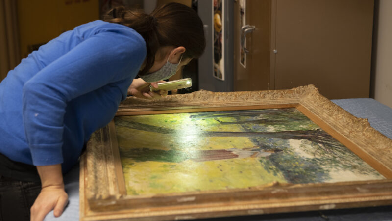 Wi=oman dressed in dark blue leaning over a painting, examining it closely with a magnifying glass.