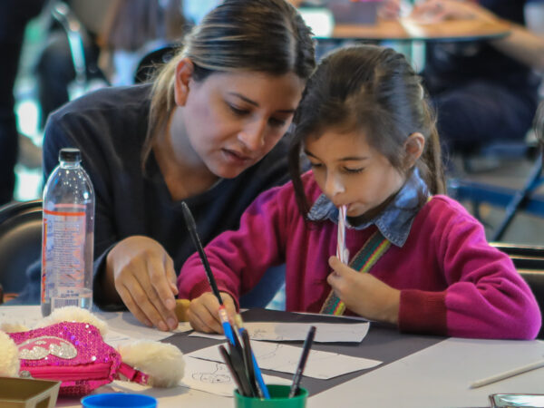 Woman with dark hair and dressed in a black top, leans toward a dark-haired girl, dressed in pink, holding a white marker to her face and looking at a piece of paper on hte table in front of them. The woman's hand is next to the child's. indicating something on the paper. A green plastic basket with pencils and other art supplies are on the table.