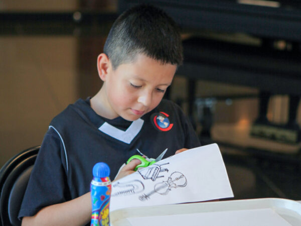 Young boy with crewcut dark hair, dressed in black t-shirt with a white V at the neckline, sits at a table and cuts musical instruments out of a white paper.