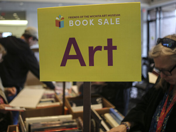 Yellow sign in the foreground with text Friends on Wichita Art Museum Book Sale, ART on a stick above boxes of books on a table. Two women in either side of the table looking at the boxes of books.