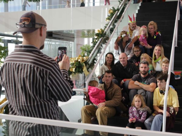 Group of people sitting on the stairs, with one person standing with their back to the camera, holding a cell phone and taking a photograph of the group.