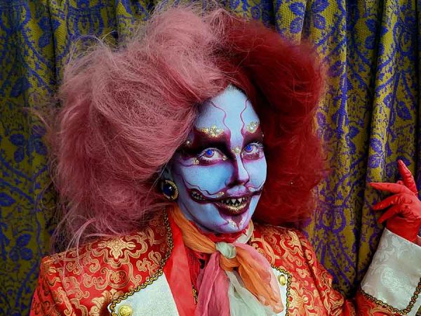 Person dressed as a clown with puffy orange hair, white face and dressed in an orange print jacket with wide white lapels.