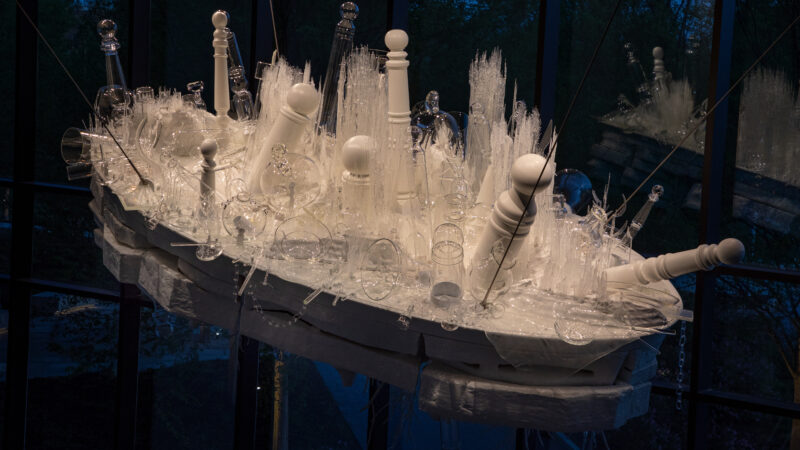 Hanging sculpture of clear glass and wood feating barber poles and decpictions of rock formations of white wood and clear glass depictions of prairie grasses, chain, vases and other objects.