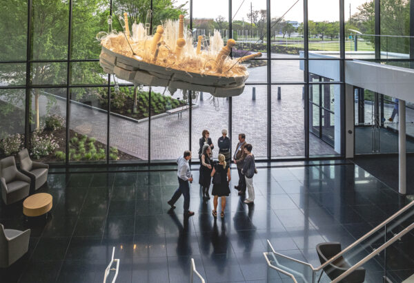 Group of 7 people standing in a semicircle below a hanging sculpture of clear glass objects and wooden poles on a white wooden base by artist Beth Lipman. The sculpture hangs in front of a wall of windows and overlooking a red brick driveway and walkway. The floor is polished black granite. The entance to a stairway with white rales is visible in the foreground.