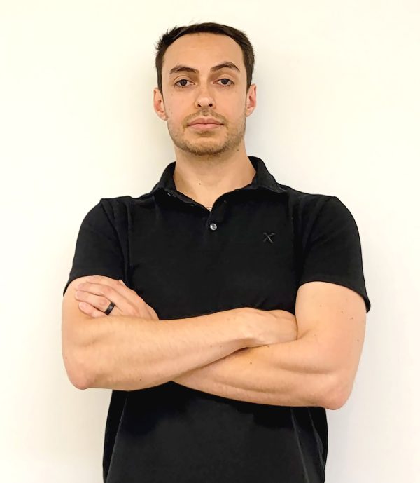 Head and shoulders photograph of a man with dark hair, dressed in a black polo-style shirt, with his arms crossed in front of his body