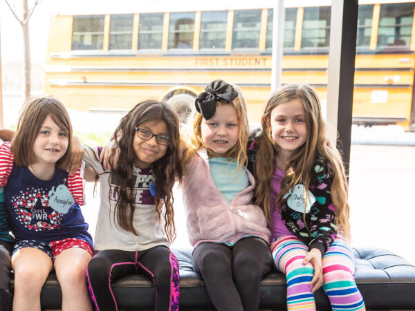 Five elementary-age students sitting on a black bench in the museum's Boeing Foyer with their arms around each other and smiles on their faces in front of the windows with a bus outside parked in the Lattner and Walker Family Plaza