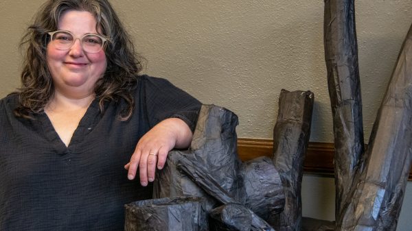 A woman with long, wavy dark hair and wearing glasses looks at the camera while standing next to elements for her art installation
