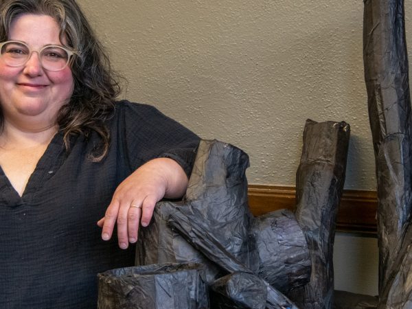 A woman with long, wavy dark hair and wearing glasses looks at the camera while standing next to elements for her art installation