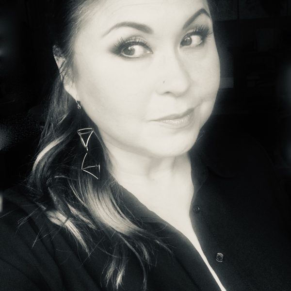 Black and white photo of a woman seen in head and shoulders slightly turned to left. She has dark hair pulled back from her face and is dressed in a dark, zip-front top.