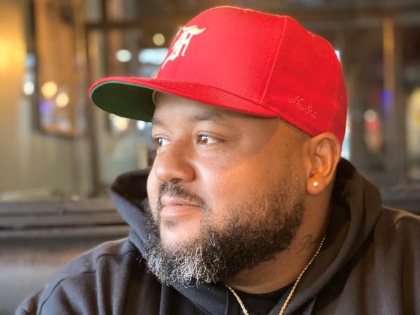 Photo of a Black man with a mustache and goatee wearing a red baseball hat and a black hoodie with a stud on his left ear and gold chain with a circular photo attached to it