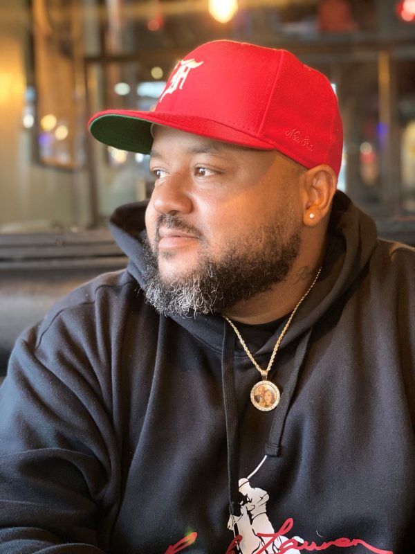 Photo of a Black man with a mustache and goatee wearing a red baseball hat and a black hoodie with a stud on his left ear and gold chain with a circular photo attached to it