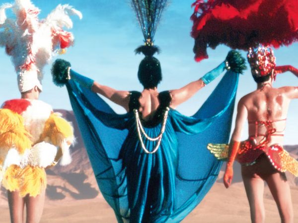 Three men in colorful costumes of yellow, blue and red stand together with their backs toward the camera and look out over the desert