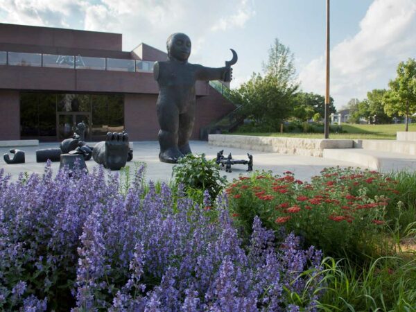 Garden photo with purple flowers in the lower left corner and red flowers on the right size. In the mddle of the image is Tom Otterness sculpture 