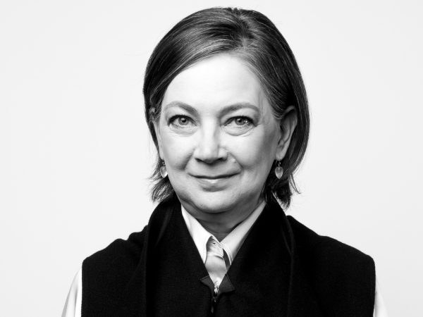 Photo of a woman with short brown hair wearing earrings, a white, long-sleeve shirt and a dark folded jacket over it