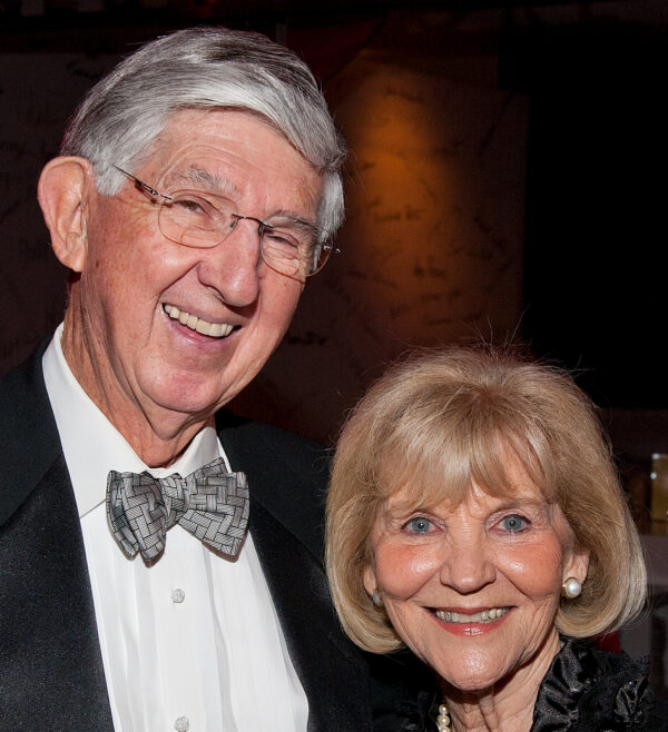 White man with grey hair, glasses and wearing a tuxedo stands next to a white woman with blonde hair, blue eyes wearing a black evening gown