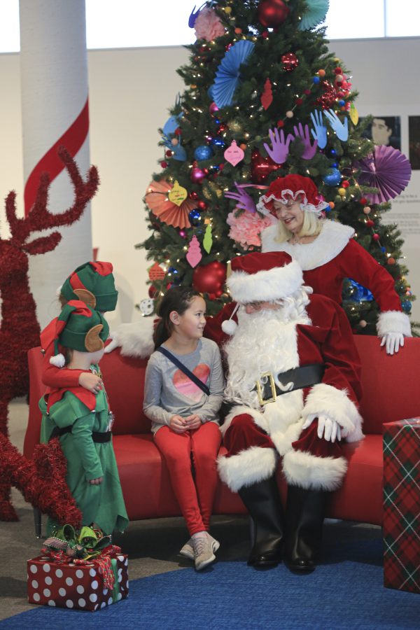 Santa Claus sitting on red bench with Mrs. Claus standing near him. Christmas tree in background. Child and elves are with Santa.