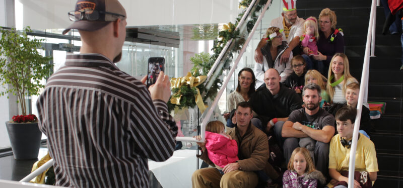 Group of people sitting on the stairs, with one person standing with their back to the camera, holding a cell phone and taking a photograph of the group.