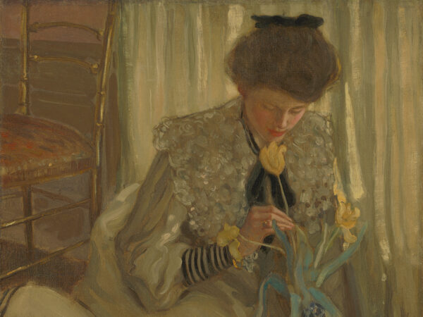 A young woman is seated on the floor leaning forward to admire one of three yellow tulips that extend from a blue and white Delftware bulb vase.
