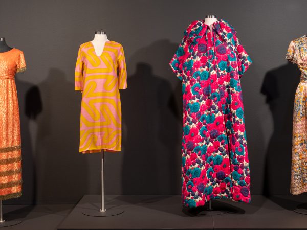 Installation image of four colorful dresses hanging on display forms: the first short-sleeve dress is long, with orange and gold; the second long-sleeve dress is short with an orange and pink pattern; the third short-sleeve jacket dress has a purple, teal, white and pink design; the fourth short-sleeve, v-neck dress has a shimmery pattern in pastel colors
