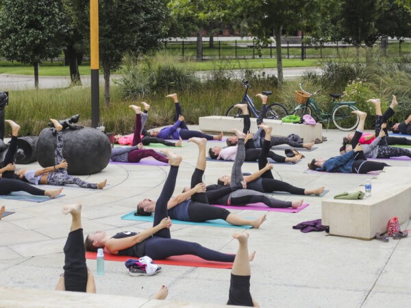 Exterior photo of the Jayne Milburn Sculpture Plaza where nearly two-dozen people are laying on the concrete plaza on colorful mats with their left legs stretched up in the air in a yoga pose
