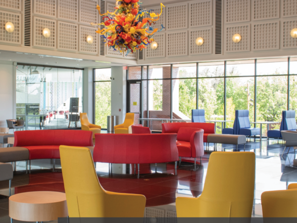View of the Farha Great Hall with the yellow, orange and blue Chihuly Chandelier, modern style yellow chairs in the foreground and a moden orange sofa in the background, framed by windows.